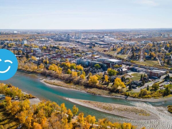 An aerial view of a vibrant riverside city in autumn, showcasing colorful trees along the riverbank and urban development in the background. A blue smiley icon is positioned in the corner, adding a cheerful touch to the scenery.