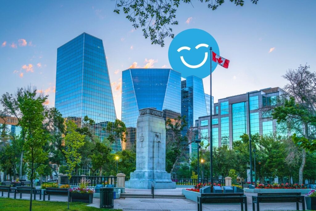 A city park features modern glass buildings in the background and a monument in the foreground, surrounded by trees and flowers. A Canadian flag is flying nearby, adding a patriotic touch to the serene urban landscape.