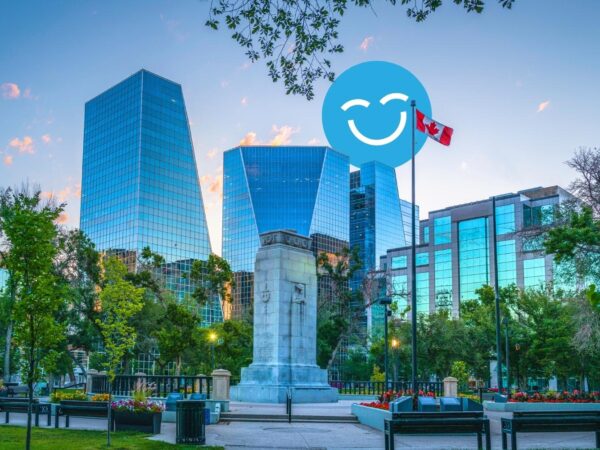 A city park features modern glass buildings in the background and a monument in the foreground, surrounded by trees and flowers. A Canadian flag is flying nearby, adding a patriotic touch to the serene urban landscape.