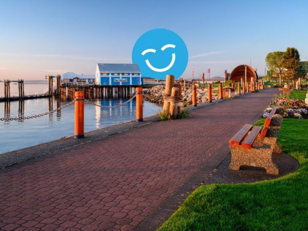 A scenic waterfront path lined with benches, leading to colorful buildings and docks under a clear blue sky. The calm water reflects the surroundings, creating a tranquil atmosphere.