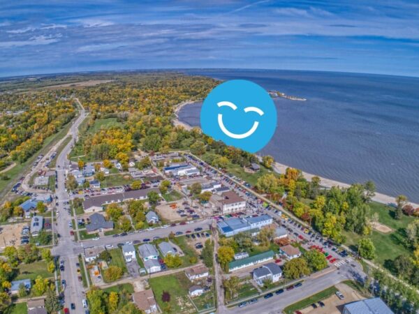 Aerial view of a small town by the water, surrounded by trees featuring autumn colors. A smiling face icon overlays the image, adding a cheerful element to the landscape.