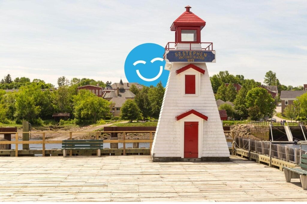 A white and red lighthouse stands on a wooden pier, surrounded by green trees and buildings in the background, under a clear blue sky with a cheerful smiley face graphic overlay.