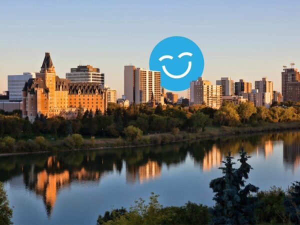 A scenic view of a city skyline at sunset, featuring modern buildings and a historic structure reflected in the calm river. Lush trees frame the foreground, enhancing the tranquil atmosphere. A cheerful blue smiley icon is superimposed on the image.