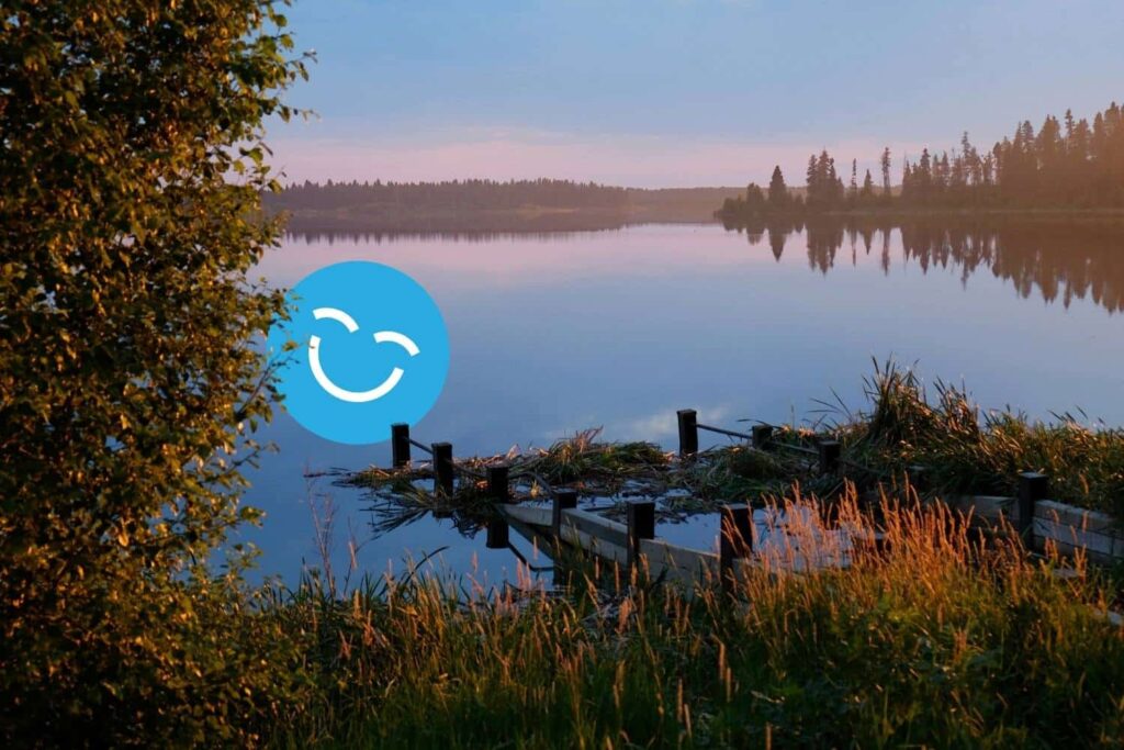 A tranquil lake scene at dusk, featuring calm water reflecting the sky and surrounding trees. In the foreground, a wooden dock extends into the water, framed by lush greenery and golden grasses. A cheerful smiley face icon is superimposed on the image.