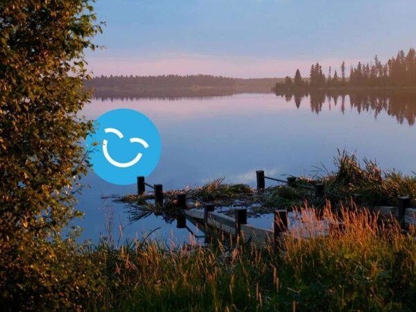 A tranquil lake scene at dusk, featuring calm water reflecting the sky and surrounding trees. In the foreground, a wooden dock extends into the water, framed by lush greenery and golden grasses. A cheerful smiley face icon is superimposed on the image.