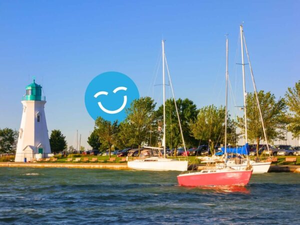 A scenic view of a lighthouse beside calm waters, with a red sailboat anchored nearby. Lush green trees line the shore under a clear blue sky, featuring a smiling icon in the background.