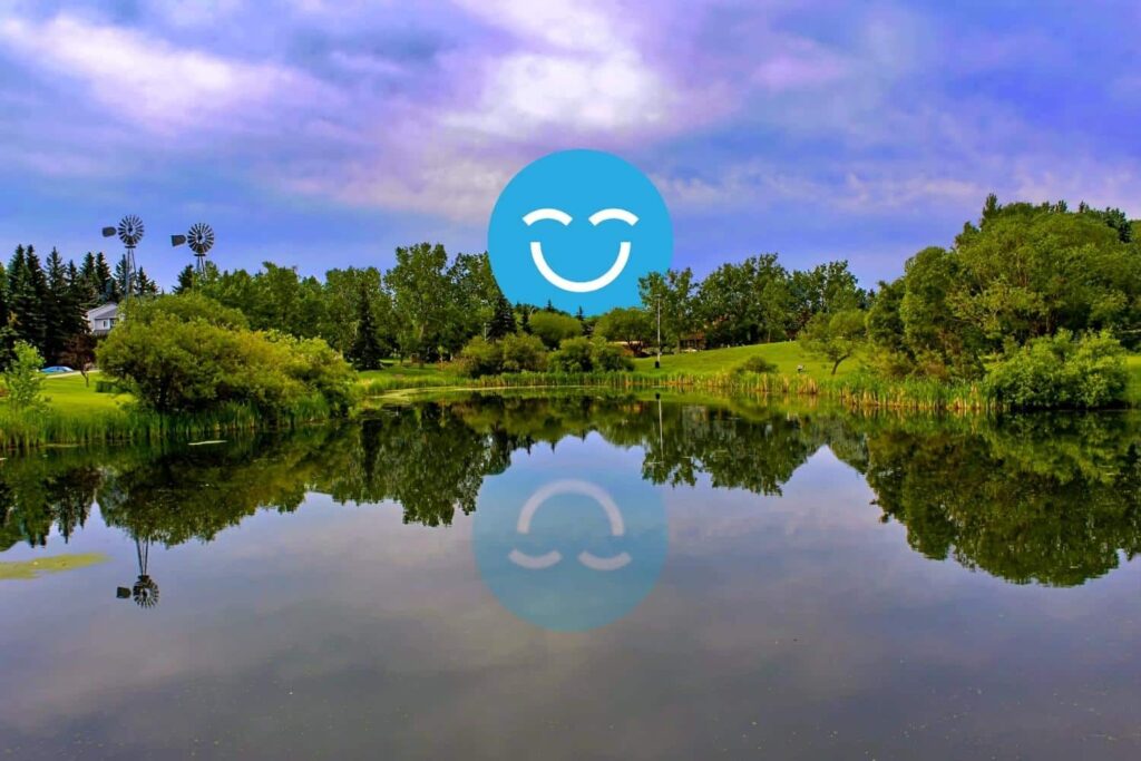 Scenic lakeside view with lush greenery and trees reflecting in calm water, under a colorful sky. A blue smiley face with closed eyes is superimposed on the scene.