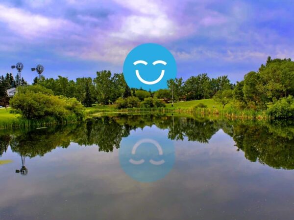 Scenic lakeside view with lush greenery and trees reflecting in calm water, under a colorful sky. A blue smiley face with closed eyes is superimposed on the scene.