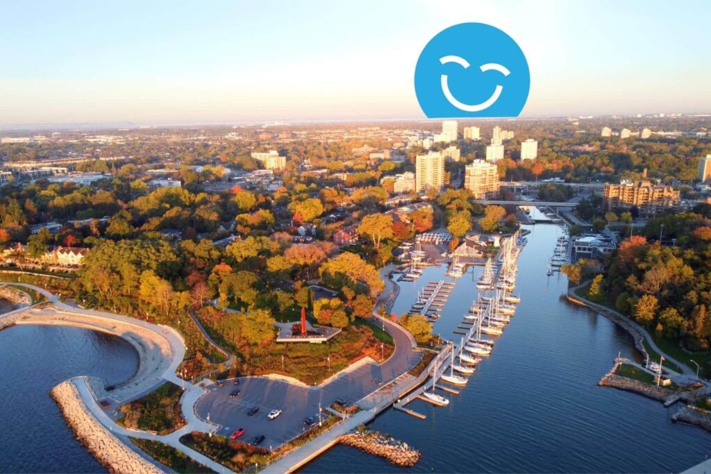 Vue aérienne d'une ville côtière au coucher du soleil, avec une marina où sont amarrés des bateaux, une végétation luxuriante et un paysage urbain en arrière-plan. Une icône bleue souriante superposée se trouve en haut.