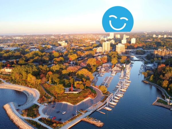 Aerial view of a coastal city during sunset, featuring a marina with docked boats, lush greenery, and cityscape in the background. An overlaid smiling blue face icon is at the top.