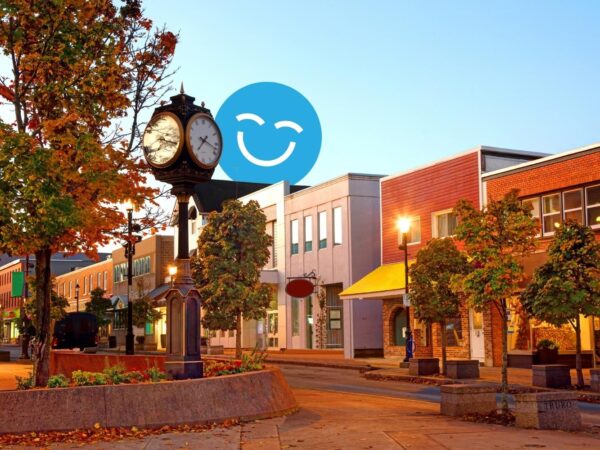 A quaint town square at sunset with a vintage clock, trees, and lit streetlamps lining the street. A smiling blue emoji is overlaid in the sky.