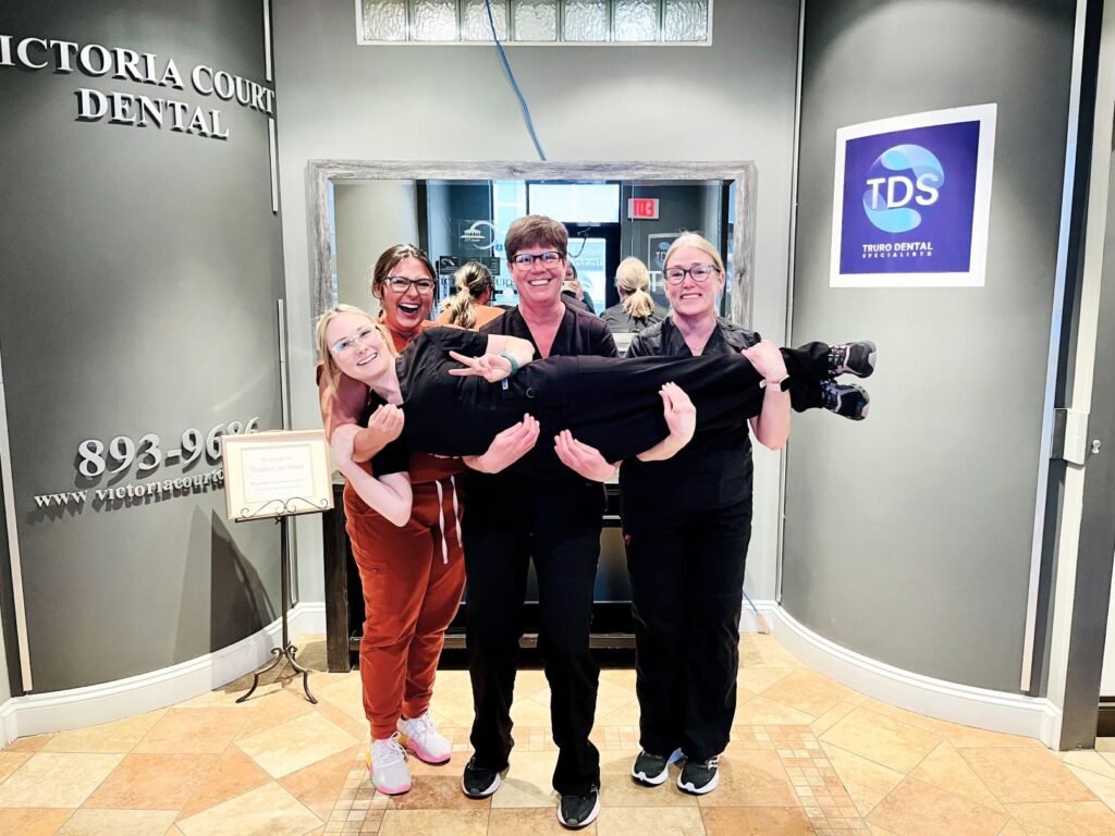 A group of four dental staff members playfully poses in a dental office, with one member being playfully lifted off the ground. The atmosphere is cheerful and friendly, highlighting teamwork and camaraderie. A blue smiley face, known as the 123Dentist Smiley, adds a touch of fun to the scene.