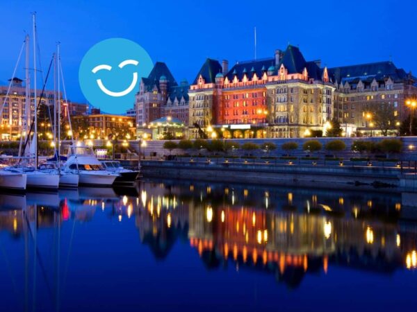 Waterfront view of a lit-up, grand building at dusk, reflecting in the calm water, with docked boats on the left and a smiling blue emoji in the sky.