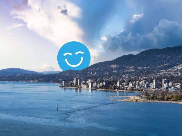 Coastal city skyline at dusk with mountains in the background and a large smiling blue face icon in the sky.