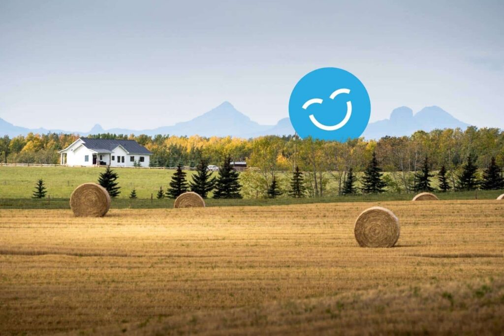 A rural landscape with golden fields, large hay bales, and evergreen trees. A white house sits in the background with mountains visible under a partly cloudy sky. A large, blue smiley face icon is overlaid in the sky.