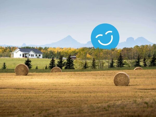 A rural landscape with golden fields, large hay bales, and evergreen trees. A white house sits in the background with mountains visible under a partly cloudy sky. A large, blue smiley face icon is overlaid in the sky.