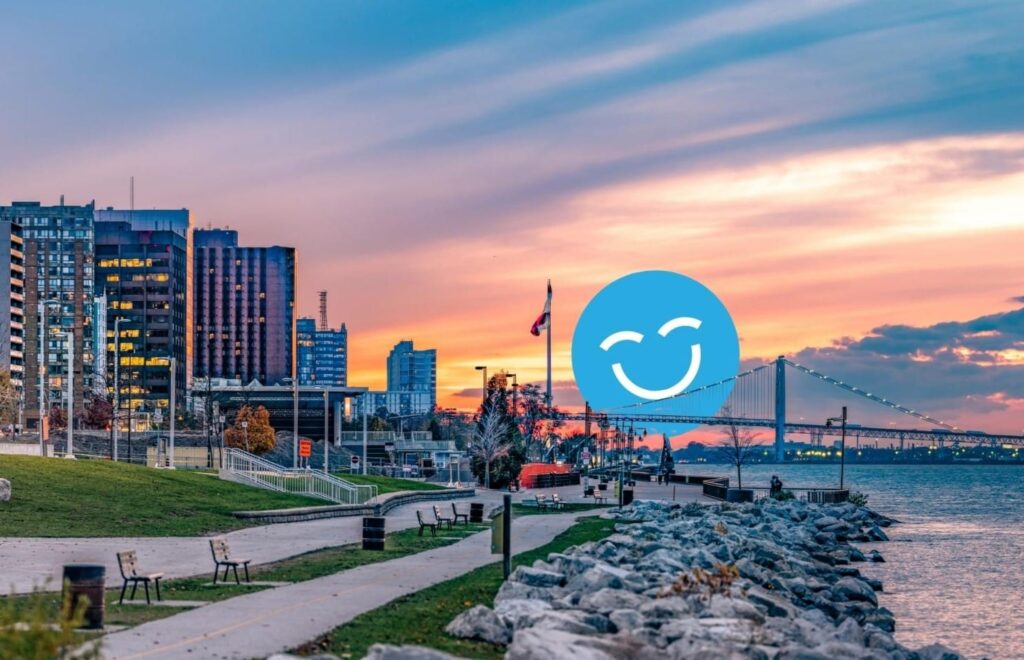 Urban waterfront scene with modern buildings, a bridge, and a sunset sky. A path lined with benches follows the water's edge, and a large blue smiling emoji is overlaid in the center.
