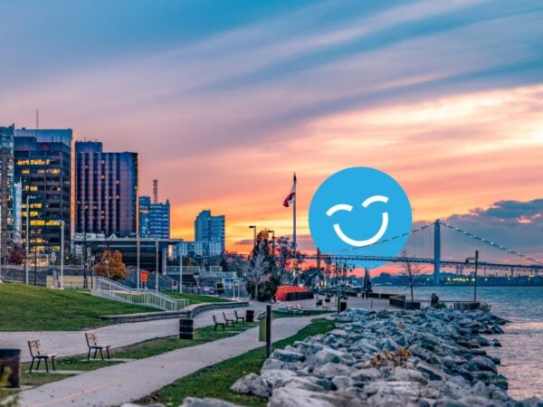 Urban waterfront scene with modern buildings, a bridge, and a sunset sky. A path lined with benches follows the water's edge, and a large blue smiling emoji is overlaid in the center.