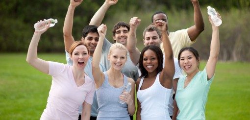 A diverse group of eight smiling individuals standing outdoors, cheerfully raising their arms and holding water bottles, celebrating together in a park.