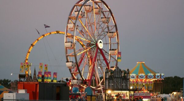 A vibrant carnival scene at dusk, featuring a large illuminated Ferris wheel, a colorful carousel, and various amusement rides with bright lights and festive decorations, creating a lively atmosphere.