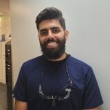 A smiling man with a beard and glasses hangs out in a medical office, wearing a dark blue scrub top. The background shows light-colored cabinetry and a modern interior.