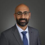 A smiling man with a beard and glasses is dressed in a formal suit and tie. He stands against a neutral gray background, exuding professionalism and approachability.