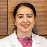 A smiling woman in a white lab coat stands against a wooden background, exuding a professional and approachable demeanor.