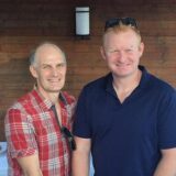 Two men stand smiling together outdoors. One man wears a red plaid shirt, while the other is dressed in a navy blue polo shirt. They appear to be in a social setting with wooden walls in the background.