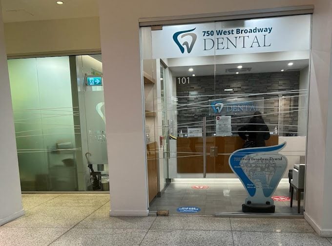 A modern dental clinic entrance featuring glass doors, a sleek reception area with a wooden desk, and the sign "750 West Broadway Dental." The interior is well-lit and showcases a professional atmosphere for patients.
