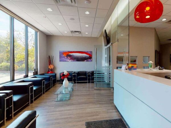A modern waiting room featuring black chairs, a glass coffee table, and a reception area with a white counter. Large windows provide natural light, and a piece of abstract art is displayed on the wall. The overall atmosphere is inviting and spacious.