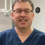 A smiling dental professional wearing blue scrubs and glasses stands in a dental office setting, with dental equipment and a clock visible in the background.