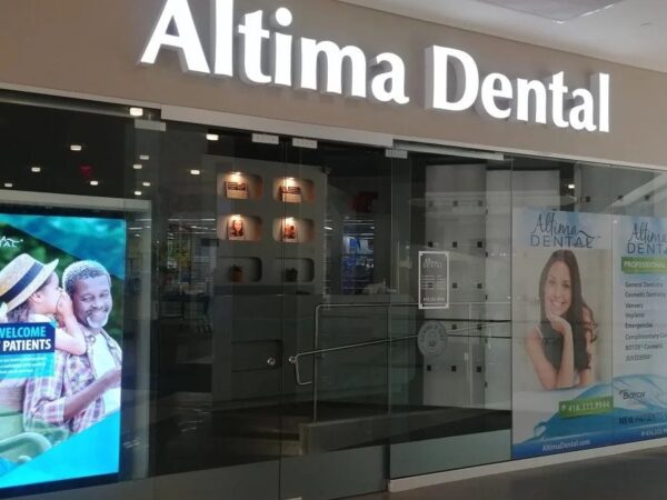Exterior view of Altima Dental office, featuring a large sign with the clinic's name. Two promotional posters display smiling patients and encourage new patient registrations, set in a modern shopping environment.