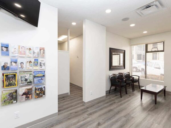 A modern waiting area features a wall with magazines, a table, and four chairs. The space is bright with natural lighting from a window, showcasing a clean and minimalist design with light-colored walls and wooden flooring.