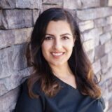 A smiling woman with long, dark hair is wearing a black top and leaning against a textured stone wall.