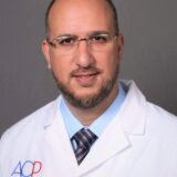 A professional man wearing glasses and a white lab coat with "Prosthodontist" and "ACP" embroidered on it, presenting a confident and approachable demeanor.