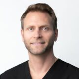 A smiling man with short hair, wearing a black scrub top, stands against a plain white background.