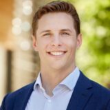 A smiling young man dressed in a blue blazer and a light blue shirt stands outdoors, with soft, blurred greenery in the background, conveying a professional and approachable demeanor.