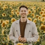 A smiling person stands in a sunflower field, holding a sunflower. The landscape features vibrant yellow sunflowers under a soft, natural light, creating a cheerful and warm atmosphere.