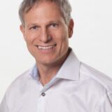 A smiling man with short, light gray hair wearing a white shirt stands against a plain white background.
