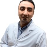 A man wearing a lab coat smiles confidently, presenting a professional appearance. He has short dark hair and a trimmed beard, standing with his arms crossed in front of a plain white background.