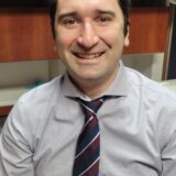 A man with dark hair smiles while seated at a desk, wearing a light purple dress shirt and a striped tie. The background features wooden cabinets, suggesting an office environment.