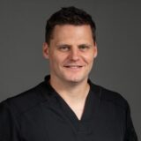A smiling individual wearing a dark scrub top poses against a neutral background.