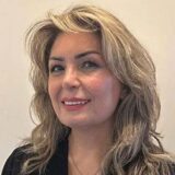 A woman with long, wavy blonde hair and a warm smile poses against a neutral background, wearing a black shirt.