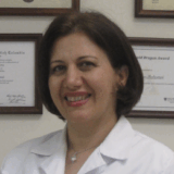A smiling woman in a white lab coat stands in front of framed certificates on a wall. She has shoulder-length dark hair and a friendly expression, suggesting a professional environment.