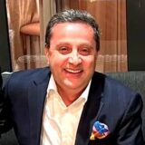 A smiling man wearing a dark suit with a white shirt, sitting on a couch. He has a colorful pocket square and is in a vibrant indoor setting.