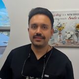 A man with a mustache is smiling while wearing a dark medical scrubs top. He is indoors, and a window with a view of trees and a wall art piece featuring flowers is visible in the background.