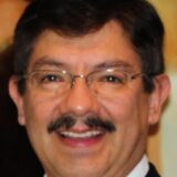 A man with short dark hair and glasses smiles warmly at the camera. He has a mustache and is wearing a suit jacket, set against a blurred background.