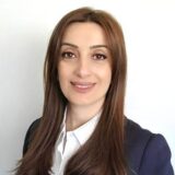 A woman with long brown hair smiles confidently while wearing a dark blazer over a white shirt. The background is neutral and unobtrusive.