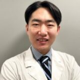 A smiling medical professional wearing a lab coat and a striped tie, posing against a neutral background.