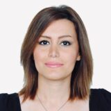 Headshot of a woman with shoulder-length dark brown hair and subtle makeup, wearing a black top. She is facing the camera with a neutral expression against a plain white background.